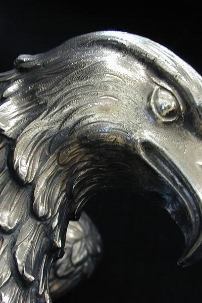 Un primer plano de la escultura Águila Gambón muestra sus detalladas plumas y su afilado pico, con un acabado suave y metálico que resalta su artesanía sobre un fondo oscuro.